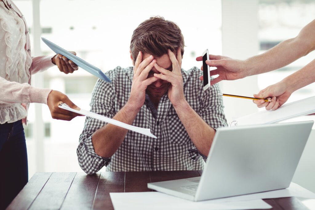 Overwhelmed man sitting at laptop with his hands over his eyes as two other people hold paperwork, a phone, and a pencil to his face.